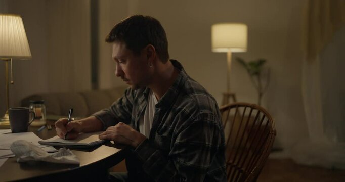 Side view of focused male writer sitting at table and writing down his ideas with pen near lamp in cozy studio. The Flow of Inspiration as a Time to Work as a Writer