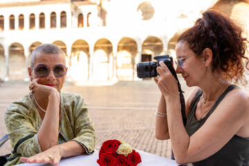 beautiful middle aged female couple having fun photographing themselves sitting at an outdoor table in a historic square