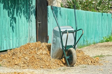 one single wheel modern new gray with green handle industrial garden iron hand cart standing on ground near clay pile in daytime outdoors near fence