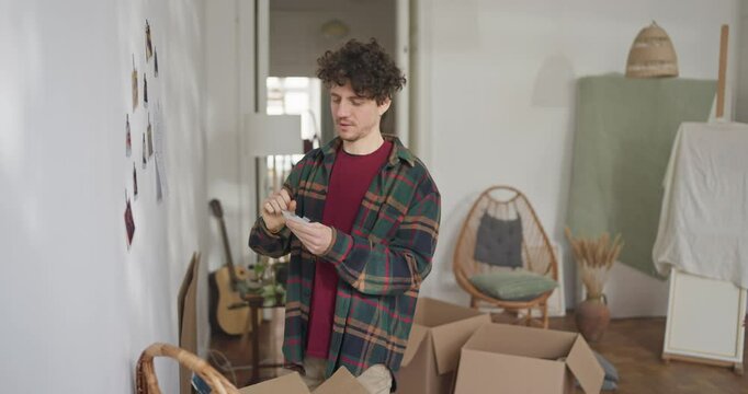 Guy with curly hair in a plaid shirt hangs photos on the wall while moving into a new bright apartment. Happy guy unpacks things in his new apartment