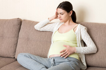 Fototapeta premium Young pregnant woman with headache sitting on sofa. Pregnancy expectation concept