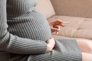 Close-up of pregnant woman is holding a cigarette sitting on sofa at home