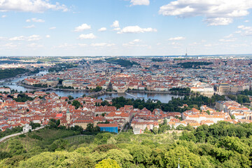Obraz premium A wide-angle view of Prague’s iconic red rooftops, historic churches, and lush green surroundings. The cityscape combines traditional architecture with glimpses of modern structures in the background