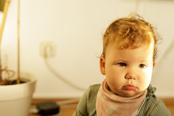 A cute baby with curly light brown hair looks at the camera with a serious expression.