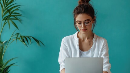 Young Caucasian woman with brown hair in a bun, wearing glasses, working on a laptop in a bright room with green walls and a plant.