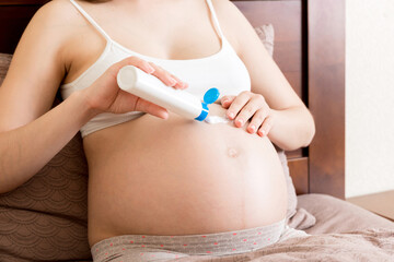 a pregnant girl sits at home on the bed and smears smile an anti-stretch mark cream on her stomach. Pregnancy, motherhood, preparation and expectation concept