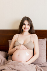 a pregnant girl sits at home on the bed and smears heart an anti-stretch mark cream on her stomach. Pregnancy, motherhood, preparation and expectation concept