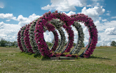 The heart-shaped arched entrances are decorated with petunias. Several identical landscape arches in the shape of a heart against a blue sky with clouds. Park design on the theme of love.