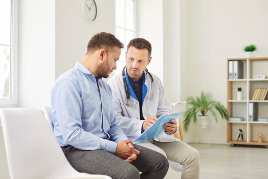 Physician or cardiologist sitting on chair with clipboard, giving consultation to male patient during checkup in medical clinic. Serious man listening carefully to treatment plan and medical advices.