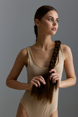 young woman with long braided hair wearing a beige sleeveless bodysuit, standing against a...