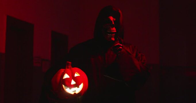 Dark macabre figure in scary Halloween costume. Sinister Death Reaper, nightmare spirit, or eerie monster disguised in cloak holds glowing Jack o lantern and shows shush shhh shut up silence gesture
