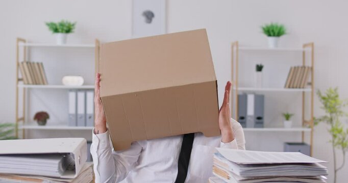 Sad company employee in eyeglasses sitting at desk, hiding his head in cardboard box, trying to escape from stress. Funny frustrated male worker taking break from work, fooling around and dancing.