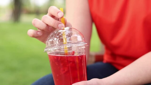 Close-up of female hands with lemonade in a disposable cup. Summer drink. Girl in a bright T-shirt sits in the park. Summer and recreation concept. Pink lemonade with raspberry and lime