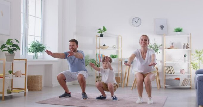 Young happy family of three with child boy doing sport exercises squatting at home. Mother, father and little son kid doing sit-ups in living room. Fitness and home training concept. 4k video.