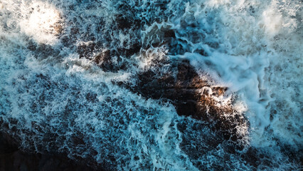 Aerial top-down view of powerful ocean waves crashing over coastal rocks, creating abstract white foam patterns. Perfect for backgrounds, marine themes, nature textures, and environmental projects.