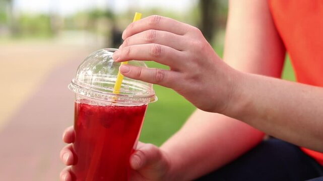 Close-up of female hands with lemonade in a disposable cup. Summer drink. Girl in a bright T-shirt sits in the park. Summer and recreation concept. Pink lemonade with raspberry and lime