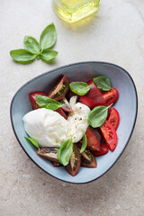 Italian caprese with buffalo mozzarella, tomatoes and fresh basil in a blue bowl, vertical shot on a beige stone background, high angle view