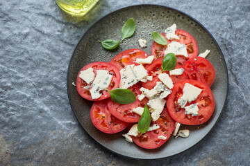 Olive-colored plate with tomato and blue cheese salad, horizontal shot on a dark-grey granite background, high angle view