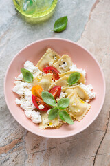 Roseate bowl with ravioli, ricotta cheese, tomatoes and fresh basil, vertical shot on a grey and roseate granite background, high angle view