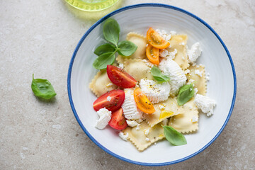 Italian ravioli served with ricotta, basil and tomatoes in a white and blue plate, horizontal shot on a beige stone background, elevated view