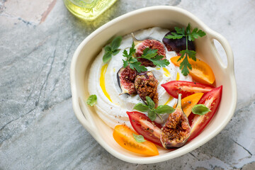 Ricotta dip with tomato wedges and torn fig fruits in a beige serving tray, horizontal shot on a grey granite background, elevated view