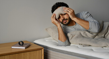young man experiencing sleep issues in modern bedroom while using eye mask for relief from discomfort and stress