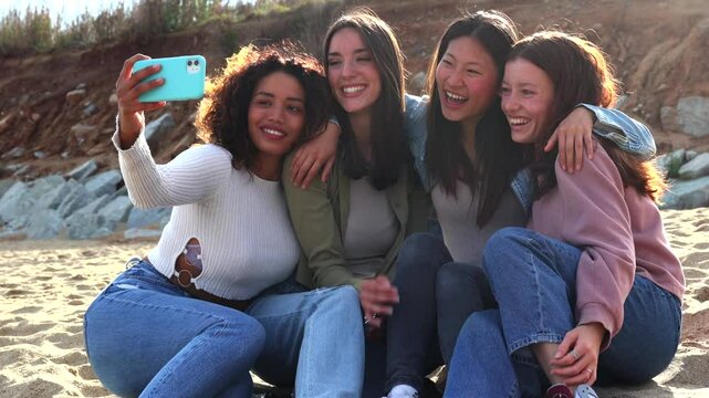 Cheerful young women creating shared memories, taking group selfie while laughing and embracing on sandy beach during sunny vacation day, capturing genuine friendship