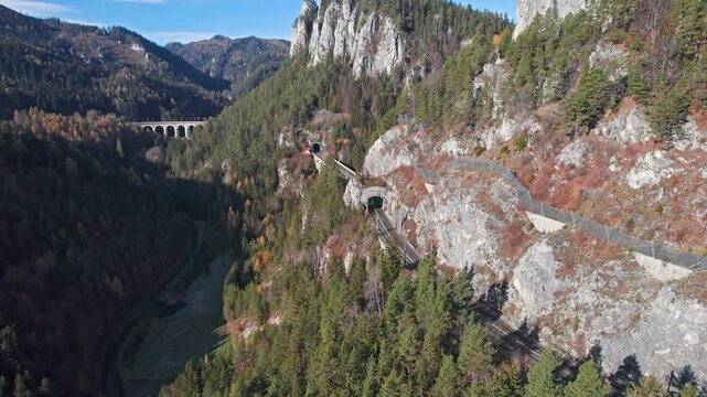 Aerial view of red passenger train rides on Krauselklause Viaduct on historical Semmering mountain railway (Semmeringbahn) surrounded by autumn forest, Lower Austria.