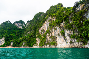 Obraz premium View of Khao Sok Lake in Krabi province, Thailand, with towering limestone cliffs covered in tropical rainforest rising above the calm surface of the lake.