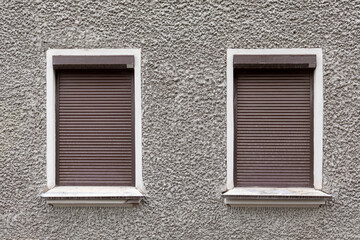 Two windows with brown shutters on a gray wall. The shutters are open, letting in some light