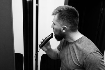 Grooming rituals in a modern setting as a man trims his beard using an electric trimmer while standing near a mirror in a contemporary space