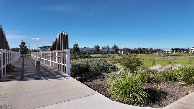Tarneit, Melbourne, Australia- a modern pedestrian footbridge spanning a landscaped green buffer zone or natural drainage corridor, with native plants, and residential housing in suburban neighborhood