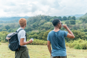 Two men standing on grassy hill, drinking and enjoying mountain scenery. Ideal for themes of travel, friendship, exploration, and outdoor lifestyle.
