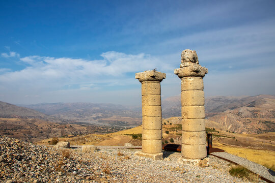 Two ancient stone columns stand on a rocky hilltop at Karakuş Tumulus in Adiyaman, Turkey. One column features a weathered eagle statue, showcasing rich archaeological heritage under clear skies.

