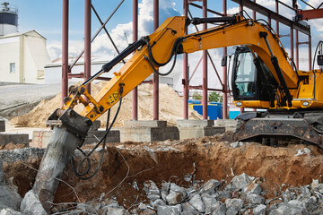 Construction site activity with excavator breaking concrete in broad daylight near industrial...