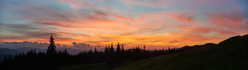 Vibrant sunset fills sky with brilliant shades of orange, pink, and purple, casting warm glow over landscape. Tall pine trees stand in silhouette against colorful horizon, with rolling hills.