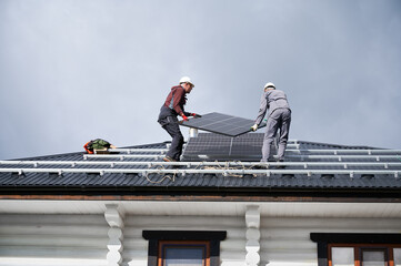 Builders installing solar panel system on roof of house. Men workers in helmets carrying photovoltaic solar module outdoors. Concept of alternative and renewable energy.