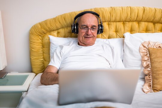Happy elderly man in pajamas using  laptop and wearing headphones while sitting in bed. - Powered by Adobe