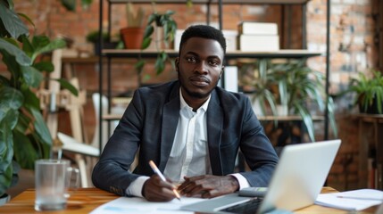 Portrait of a young male office worker sitting at a desk and writing documents, typing on a laptop, looking seriously and confidently at the camera.