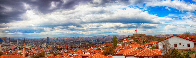 Naklejka premium Panoramic view of Ankara Castle with Turkish flag on rocky hill, surrounded by traditional red rooftops and cityscape under dramatic cloudy blue sky, showcasing Turkey’s historic and urban contrast.
