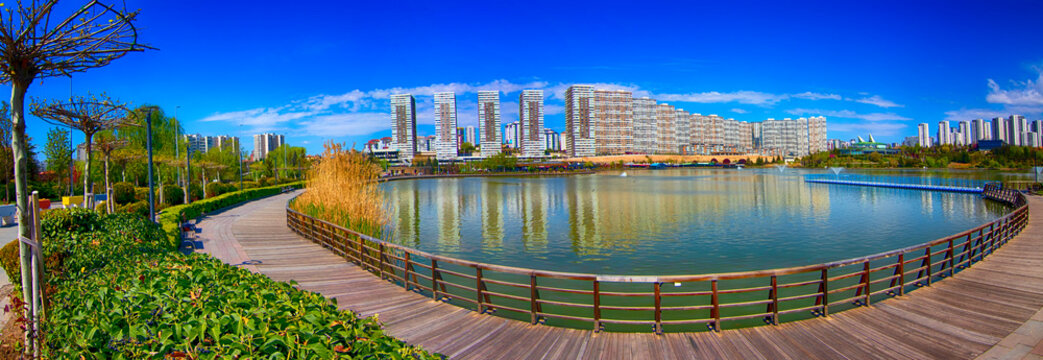 Panoramic view of Ankara Göksu Park featuring curved wooden walkway around calm lake, lush greenery, modern residential buildings, and vibrant blue sky on a sunny day in Turkey’s capital city.
- Powered by Adobe