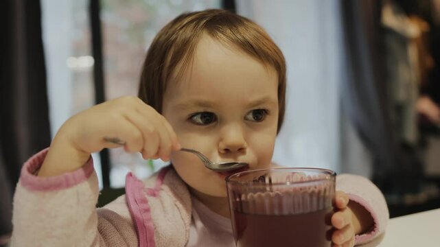 Close-up portrait of a beautiful girl drinking compote or juice with a spoon in the living room while sitting at the table and watching TV.