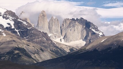 The iconic granite spires of Torres del Paine rise above glacial terrain in Chilean Patagonia, framed by snow-dusted ridges and thick clouds over a dramatic mountain landscape.