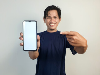 Young Asian Man Smiling While Pointing at Smartphone with Blank Screen, Casually Dressed in Blue...