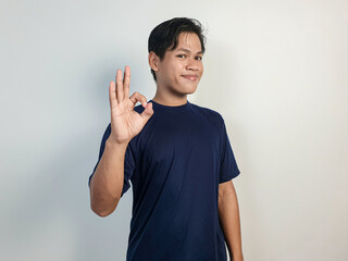 Smiling young Asian man in a blue shirt giving an OK hand gesture against a solid white background....