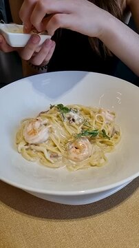 Vertical slow-motion closeup of female hand sprinkling parmesan over spaghetti with shrimp on a white plate. Captured with soft studio lighting.