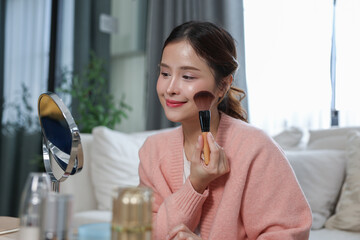 Young woman happily applying makeup blush brush on her cheek in front of mirror at home, enjoying...