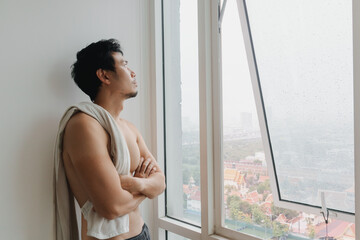 Shirtless Thai man standing by open window in city apartment