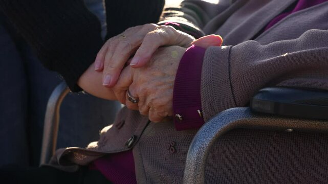 Granddaughter gently holding elderly woman’s hands in wheelchair. Close up