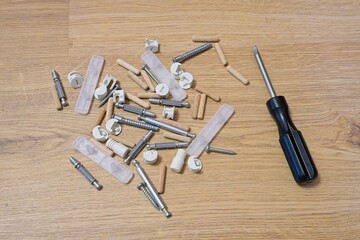 Furniture assembly hardware scattered on wooden floor, metal screws, dowels, plastic components surrounding black screwdriver